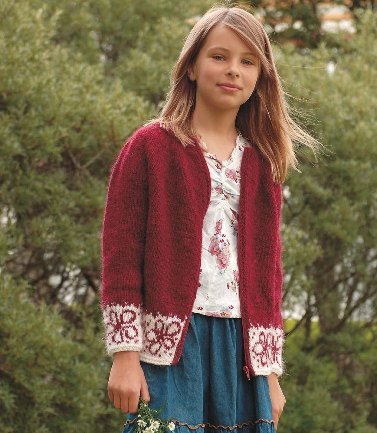 Girl in Blóm, worn open, in the pattern’s colours — a garnet red Léttlopi zip cardigan with white bands of red flowers at the hem and on the cuffs, a plain red body, sleeves and yoke, red front bands and a red zip. The floral blouse, the denim skirt and the daisies in her hand are not part of the pattern