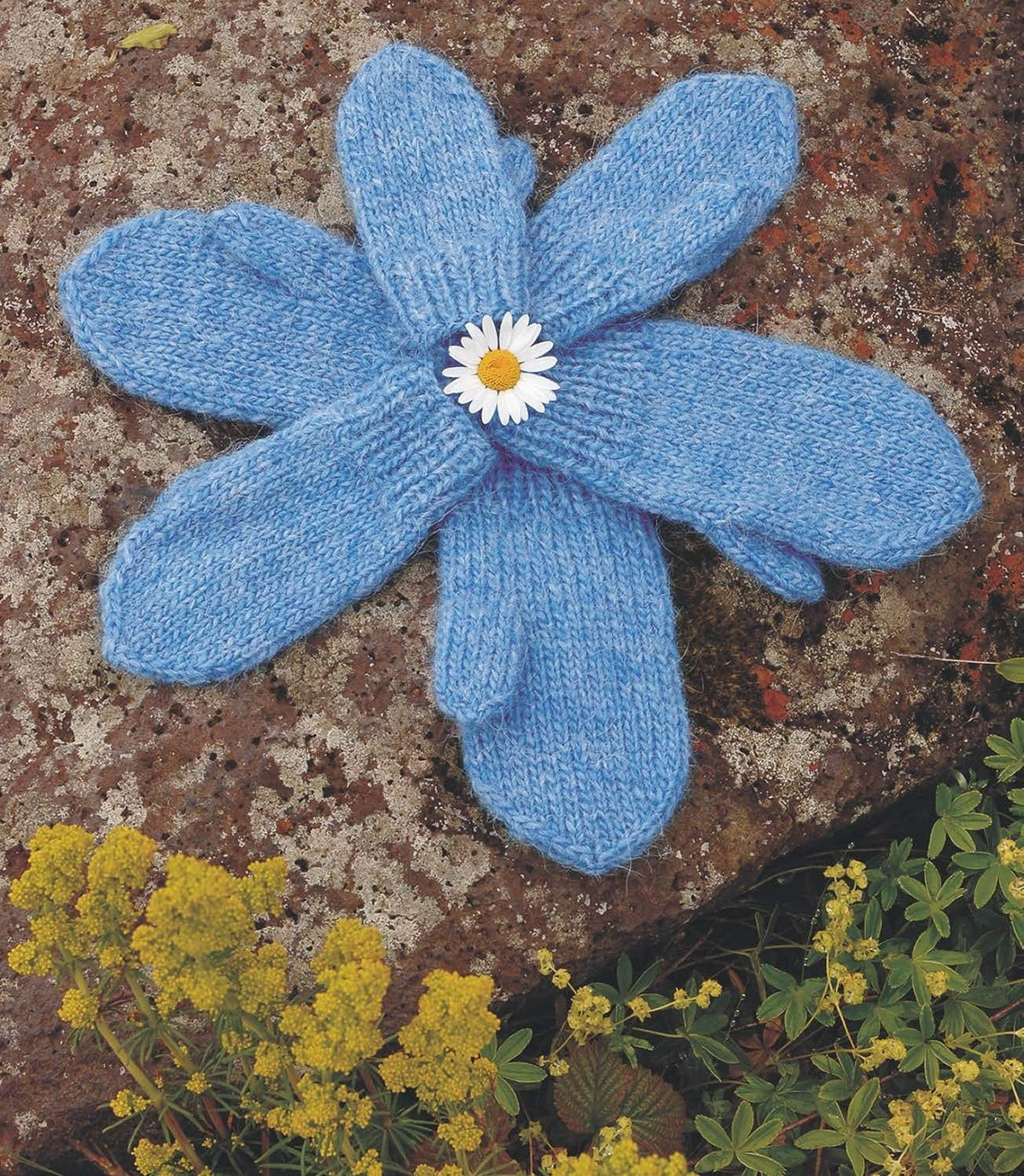 Six heaven blue Kríli mittens laid in a flower shape on a rock around a daisy