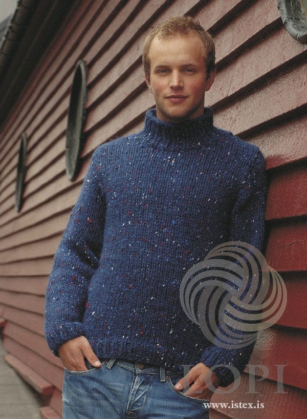 The same man against a red wooden wall in the blue version of Strax — mountain blue, a blue tweed with red, blue and white flecks, in plain stocking stitch with the polo collar all in blue, shown in the pattern’s original colour (no longer made). The jeans are not part of the pattern