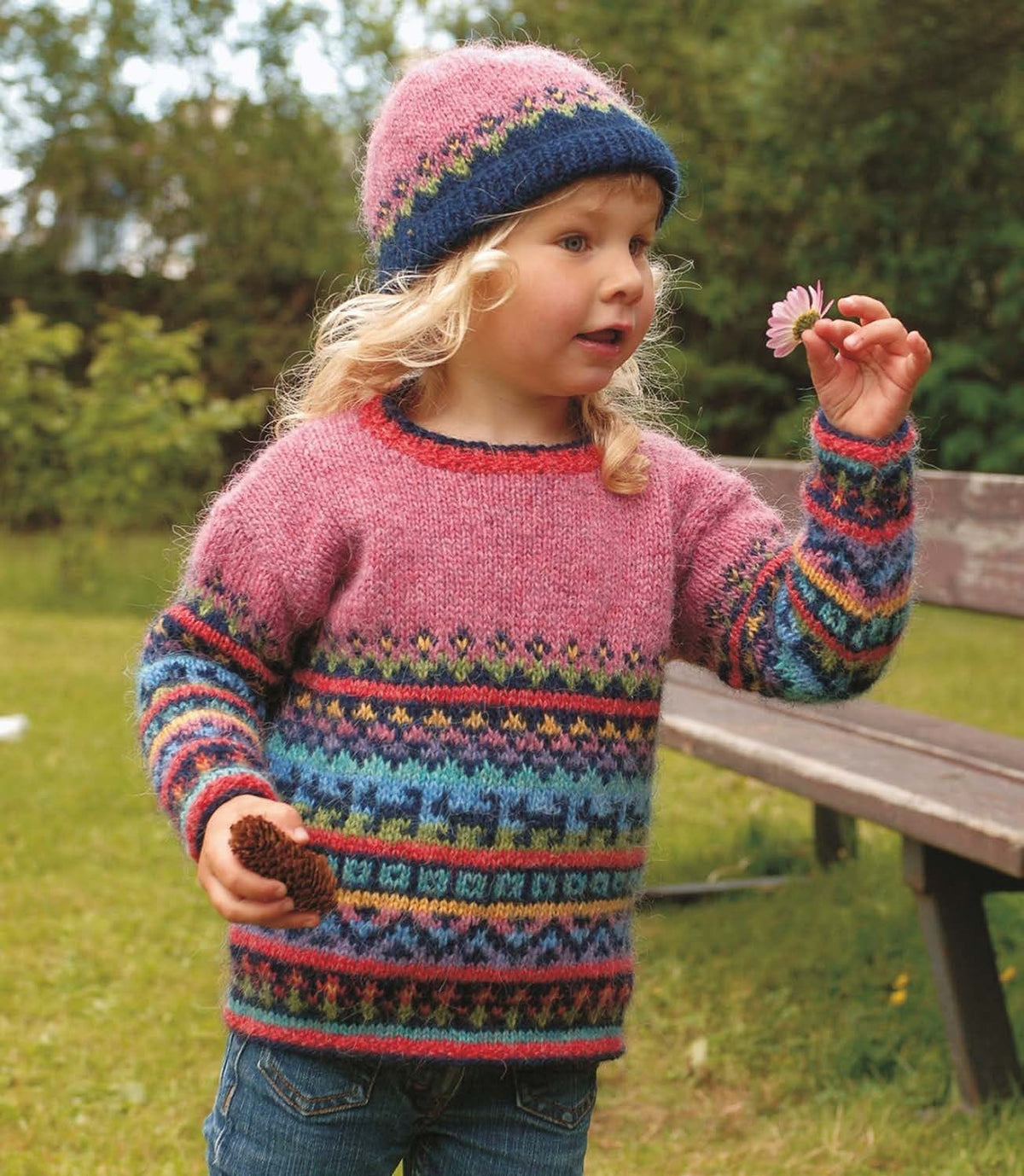 Little girl in a garden in the Týra sweater — a plain pink upper body and sleeve tops over narrow bands of flowers, zigzags, stripes and squares in navy, red, yellow, blues, green, lilac and pink, with red moss-stitch edges and a red neckband edged in navy (shown in the original colours — this kit has crimson red for the light red, mimosa for the sun yellow and milkyway for the lilac). The hat is a separate kit; the jeans are not part of the pattern