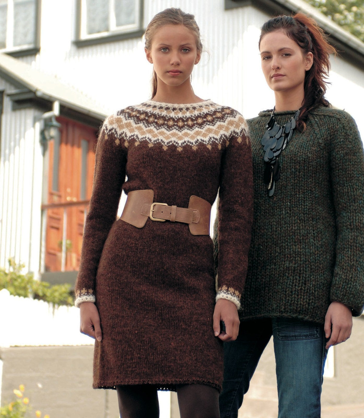 Woman in the brown Eyja sweater dress, reaching just above the knee — a reddish-brown dress with a round yoke of white and camel zigzags and diamonds, a white neck edge and white-and-camel cuff borders (shown in the original colours — this kit has chocolate heather for the brown and barley for the camel). The wide leather belt and the tights are not part of the pattern; the green sweater on the woman beside her is a different pattern