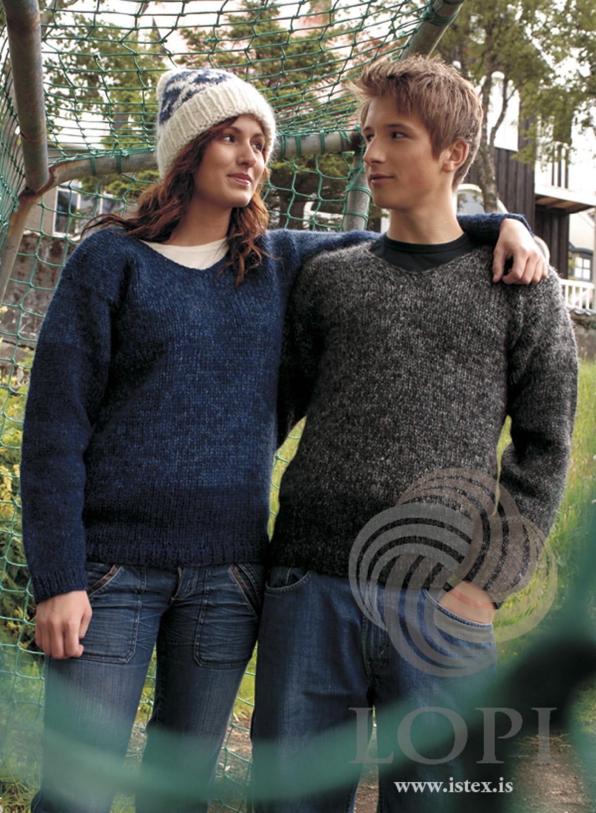 Two people in Húm by a football goal, both in the pattern’s own colours — the woman in the blue version, a blend of winter blue, navy and arctic blue with a darker navy band at the hem, the man in the grey version, a dark tweedy blend of dark grey, black heather and grey heather. Her white patterned hat, the T-shirts and the jeans are not part of the pattern