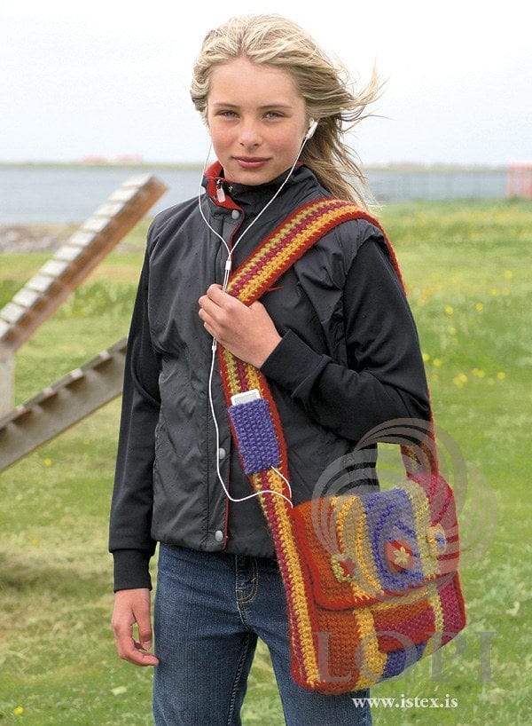 Girl wearing the Sól bag across her body — single-crochet stripes in rust orange, golden, violet, chartreuse and fuchsia with a red edge, three small squares with flowers on the flap, and a violet knitted pocket on the striped strap (shown in the original colours — this kit has space blue for the ultra violet and amber heather for the orange rust). The jacket, jeans and music player are not part of the pattern