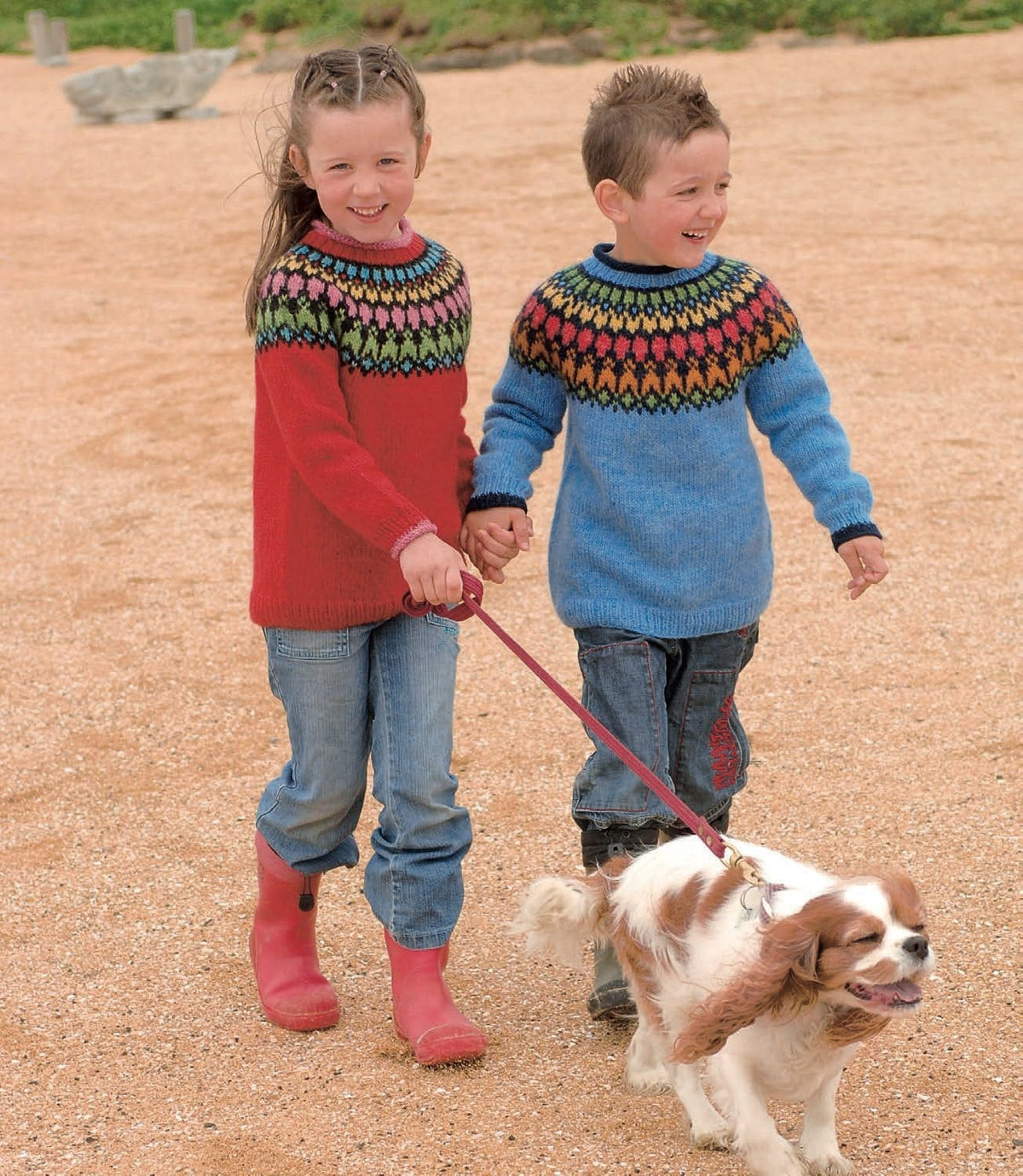 Two children holding hands on a sandy path. The boy wears the blue Þíða sweater — heaven blue with a navy round yoke of green, orange, red and yellow motifs, and navy rolled edges at the neck and cuffs (shown in the original colours — this kit has apricot for the orange, crimson red for the light red and mimosa for the sun yellow). The girl wears the red version, sold as a separate kit. The jeans, boots and dog are not part of the pattern