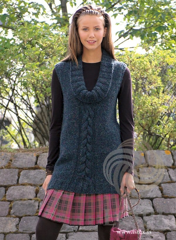 Young woman by a stone wall in U — a long sleeveless tunic with one braided cable up the centre front and a deep K2, P2 rib collar falling into a cowl, shown in the pattern’s original colour, pond heather (9736, no longer made), a dark petrol-blue heather. The black long-sleeved top, the pink plaid skirt, the dark tights, the bracelet, the hairband and the pink cabled bag are not part of the pattern