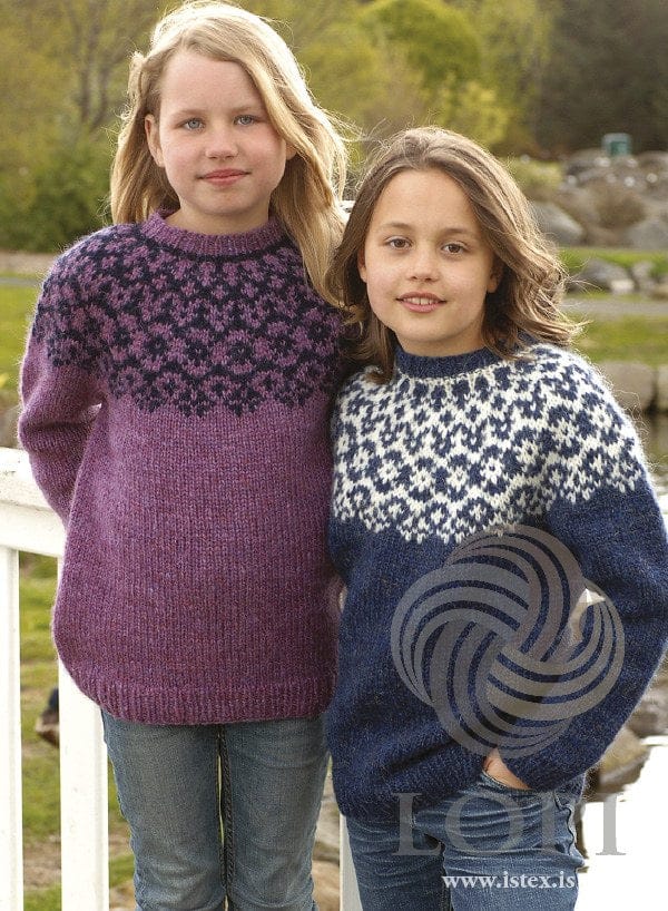 Two children at a white wooden railing in Él sweaters, both in the pattern’s original colours. On the left, the purple version — hyacinth (no longer made) with the yoke patterned in midnight blue and a dark line at the base of the neckband; on the right, the blue version — winter blue with a white yoke of small blue diamonds and flowers and a white line at the base of the neckband. Their jeans are not part of the pattern