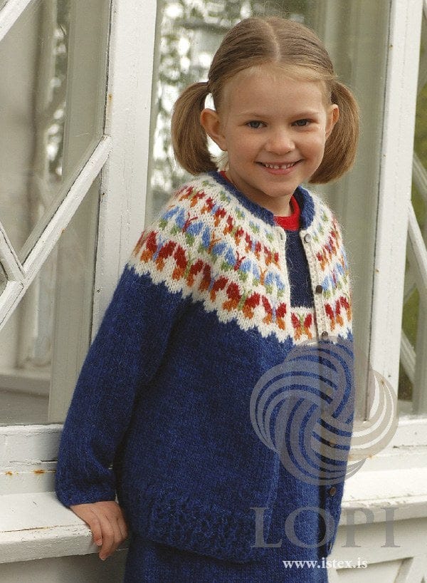 Smiling girl with pigtails in Fiðrildaslóð, the blue version — lapis blue with a white round yoke of butterflies in orange, crimson red, spring green and heaven blue, eyelet rib at the hem and a crochet border down the front, shown in the pattern’s original colours (orange, 1410, is no longer made). Her red top and the blue knitted garment under the cardigan (seen at the front opening and below the hem) are not part of the pattern