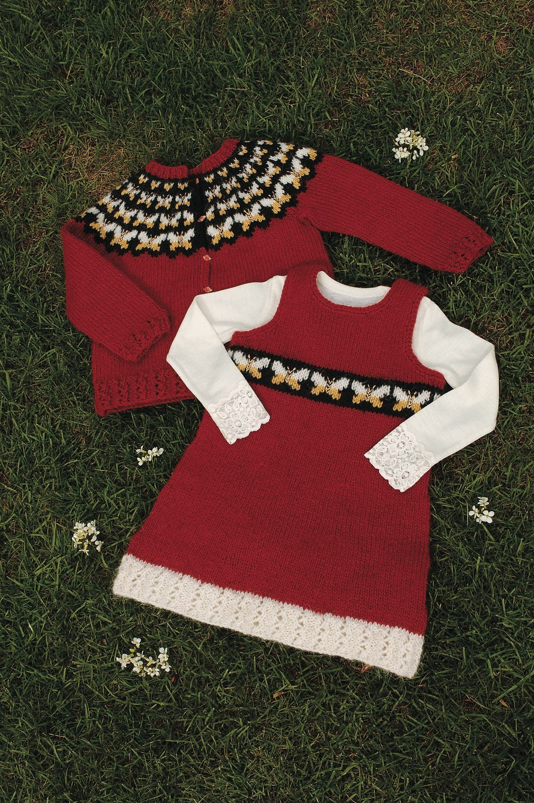 The Skotta dress in its red version, laid on the grass over a white blouse — crimson red with a white eyelet-rib hem and a black band of white and yellow butterflies. There is no photo of the blue version, which has a lapis blue skirt, a white band of crimson red and apricot butterflies (apricot replaces the pattern’s orange) and, by our reading, a navy bodice. The blouse is not part of the pattern; the red cardigan behind is Fiðrildaslóð, sold as a separate kit