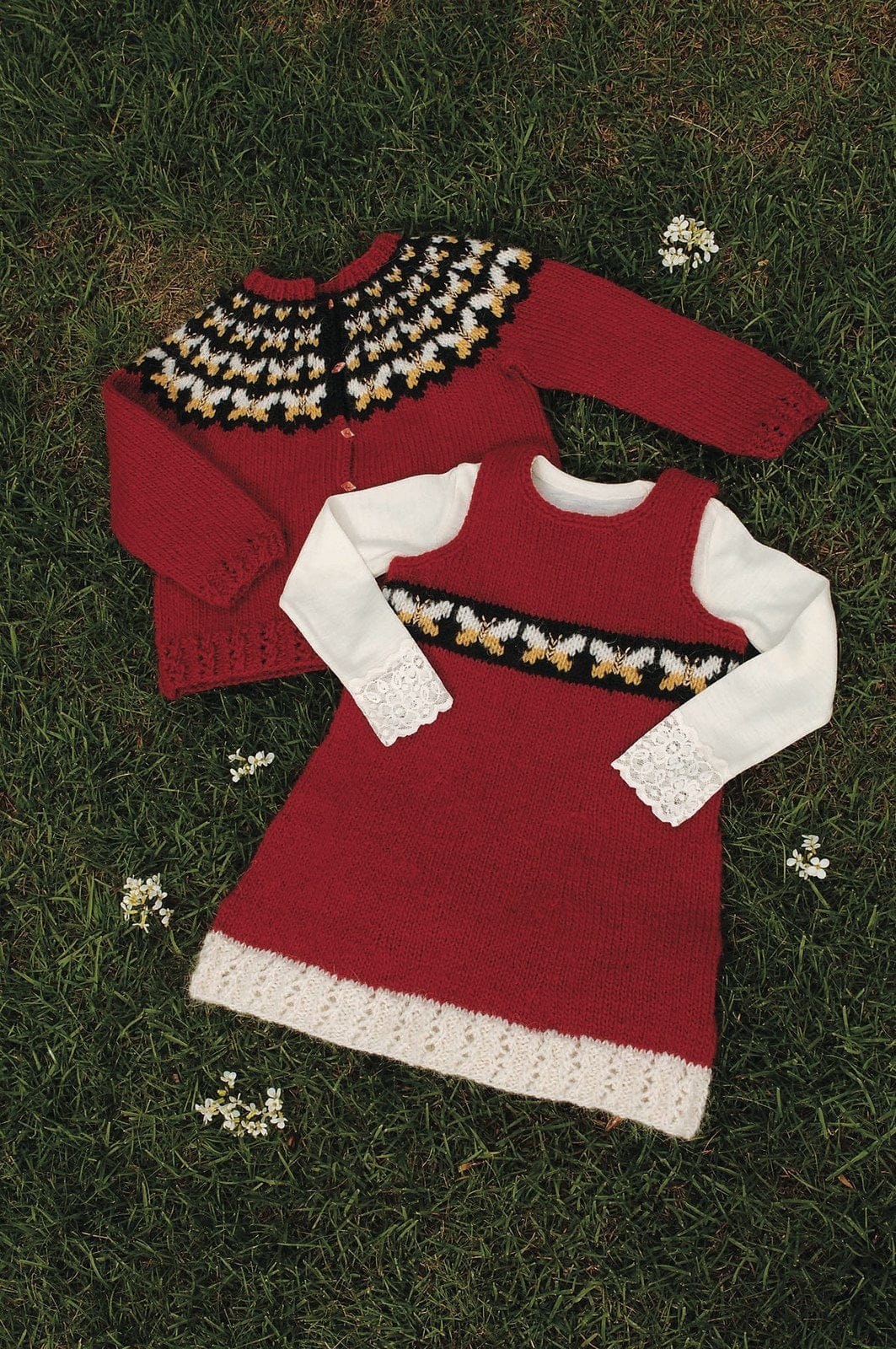 The red version of Fiðrildaslóð lying on grass — crimson red with a black round yoke of butterflies with white upper wings and yellow lower wings, small buttons down the front and eyelet rib at the hem and cuffs, in the pattern’s original colours (sun yellow, 1411, is no longer made). The Skotta leaflet (Lopi 27 no. 2) shows this photo cropped to the dress: the red dress is Skotta, a separate pattern; the dress, the white lace-cuffed top under it and the small white flowers are not part of this one