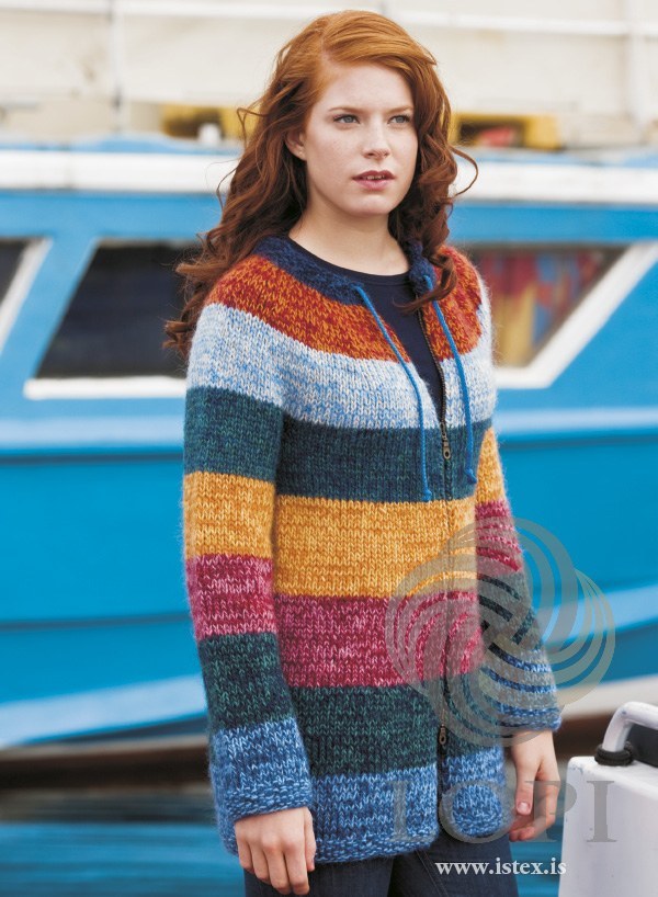 Woman in the Hamingja cardigan, zipped over the body and open at the neck — wide blended stripes of blue, dark teal, raspberry, golden and dark blue-green, and a round yoke with a pale blue-grey band, a red-orange band, a dark neckband and a bright blue knotted cord (shown in the original colours — this kit has apricot for the orange and mimosa for the sun yellow, so the golden stripe and the orange yoke band are deeper). The cord is not included; the navy top and jeans are not part of the pattern