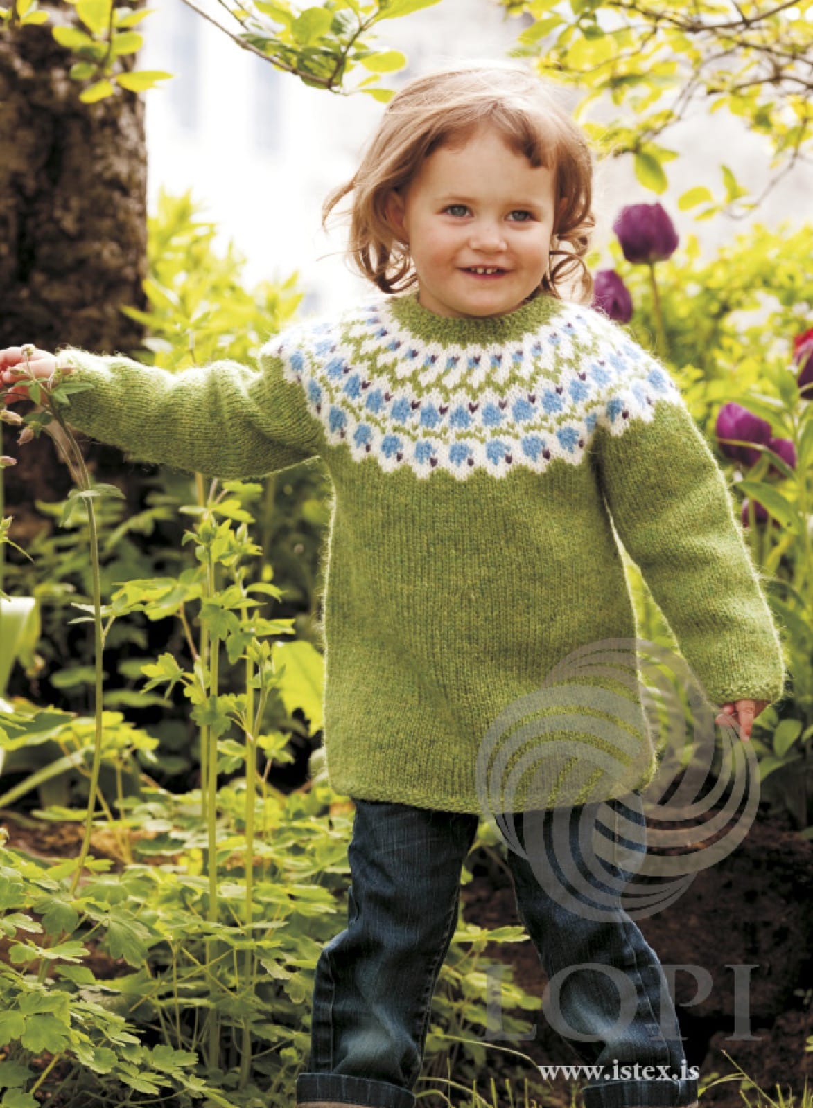 Toddler in a garden wearing Bláklukka, the green version — a spring green body and sleeves, a white yoke band with heaven blue bell flowers, small violet accents and green zigzags, and a green moss-stitch neckband, shown in the pattern’s original colours. The jeans are not part of the pattern