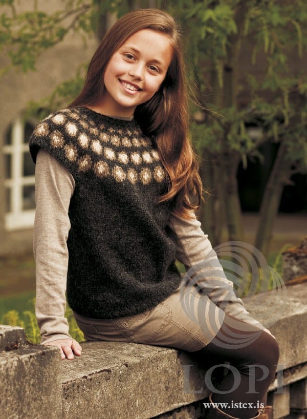 Girl sitting on a stone wall in Doppa, the natural version — black heather with rows of murky and straw dots round the yoke, short cap sleeves and a plain body, shown in the pattern’s own colours. The beige long-sleeved top, the beige shorts, the dark brown tights and the boots are not part of the pattern