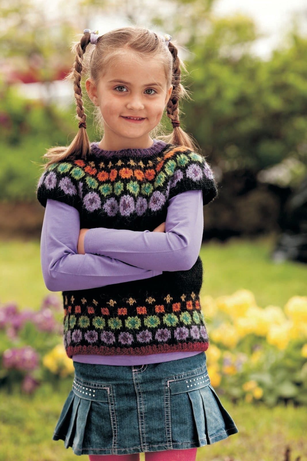 Girl in the colourful Doppa vest — a violet neck rib, a black yoke with rows of small yellow flowers, orange squares, green dots and big lilac dots, short cap sleeves, and the same dots above a garnet hem rib (shown in the original colours — this kit has mimosa for the mustard, apricot for the orange and milkyway for the lilac). The lavender top and the denim skirt are not part of the pattern