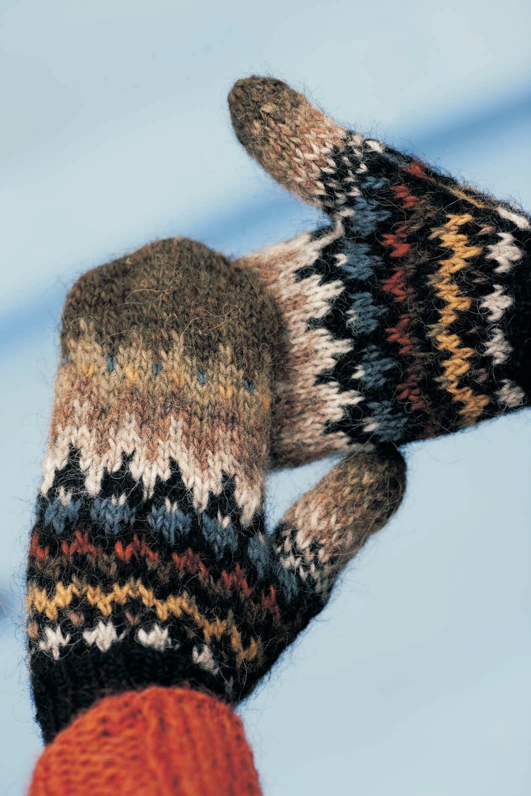 A pair of Handtak mittens in the black-moss version against a pale blue background — black cuffs, narrow bands of crosses, zigzags and dots in light ash, beige, stone blue, rust and golden mustard, rising to a solid brown-green moor top and thumb tip, shown in the pattern’s original colours (mustard is no longer made). The orange sweater cuff is not part of the pattern