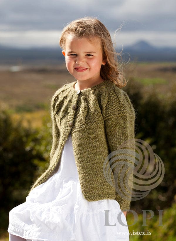 Girl outdoors in Laufey in clover green, the pattern’s green version — a yoke of twisted-stitch ribs with small leaves, garter-stitch edges and a stocking-stitch body, buttoned at the top with three buttons (not included). The white dress and the pale tights are not part of the pattern