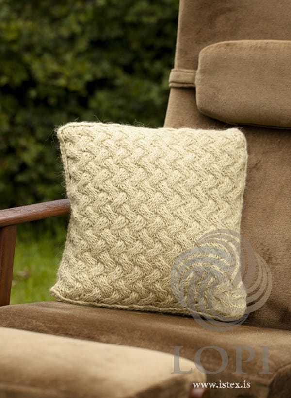 The Þari cushion on a brown armchair — a square cover with a front of interlocking basketweave cables and a braided cord round the edge, shown in the pattern’s original colour, fairy green, a pale greenish oat (no longer made). The armchair and the footstool are not part of the pattern