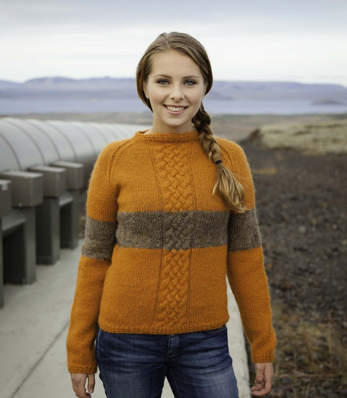 Woman in the orange Gná sweater — a raglan pullover with a braided cable up the centre front, a grey-brown band across the chest and sleeves, ribbed hem and cuffs and a narrow ribbed neck (shown in the original bright orange — this kit has apricot, a deeper, browner orange, in its place). The jeans are not part of the pattern