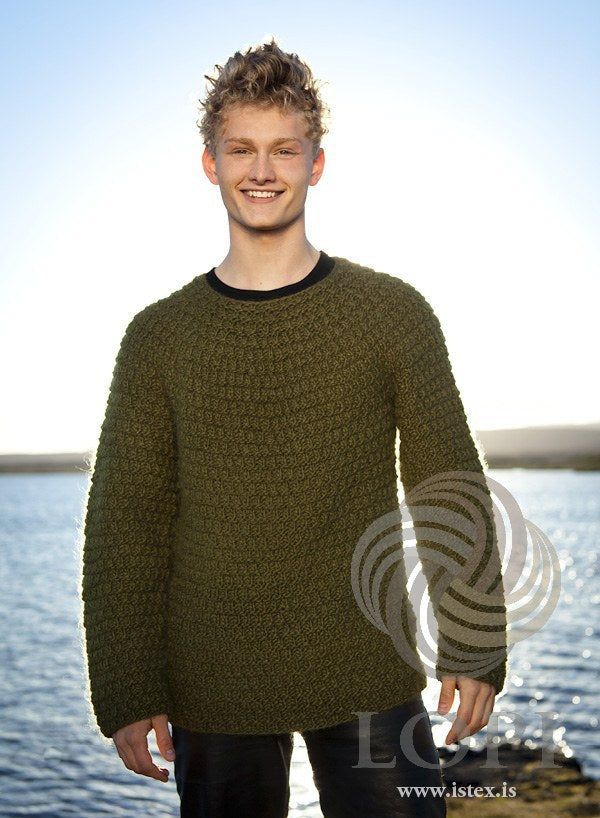 Young man by the water in the Múr pullover — an all-over texture of purl ridges and small offset blocks, a round yoke and a plain neck with no rib; the black T-shirt at the neck is not part of the pattern (shown in the original colour, green moss — this kit has highland green, a little browner)