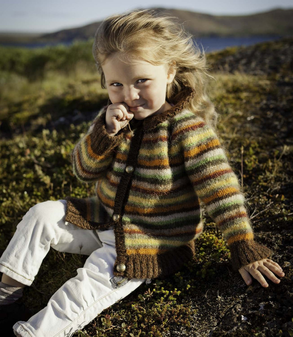 Small girl sitting on heath in the Sara cardigan — narrow stripes of orange, brick red, yellow, green, grey-green, beige and dark moss, with brown flared cuffs, a brown ribbed hem, brown front bands, round buttons and a big brown collar (shown in the original colours — this kit has chocolate heather, a darker brown, for the hazel heather, apricot for the orange and mimosa for the sun yellow). The buttons are not included; the white trousers are not part of the pattern