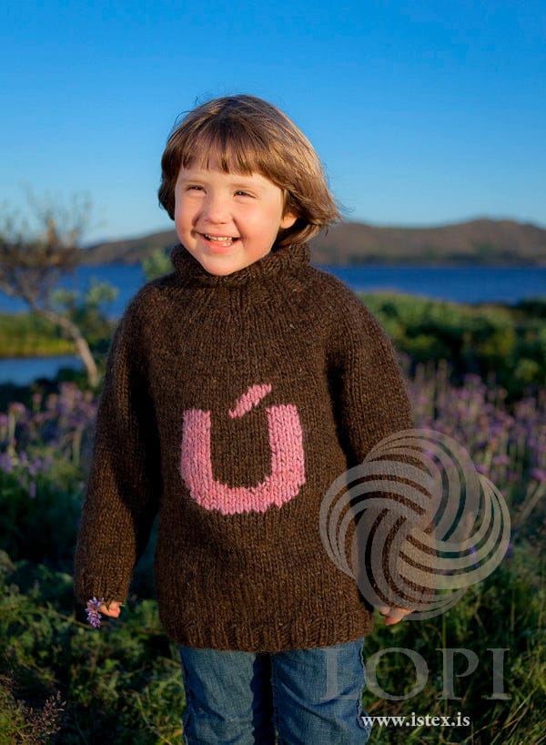 Smiling child by a lake among purple flowers in Úlfhildur, shown in the pattern’s own colours — chocolate Álafosslopi with a round yoke, a tall ribbed collar folded over and a big pink Ú sewn on the front. The jeans and the small purple flower in the child’s hand are not part of the pattern