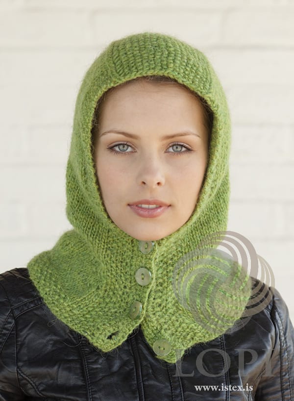 Woman wearing Hetta up as a hood, buttoned under the chin, in the pattern’s spring green heather — moss-stitch edges round the face, a moss-stitch panel over the crown and four buttons. The black leather jacket is not part of the pattern