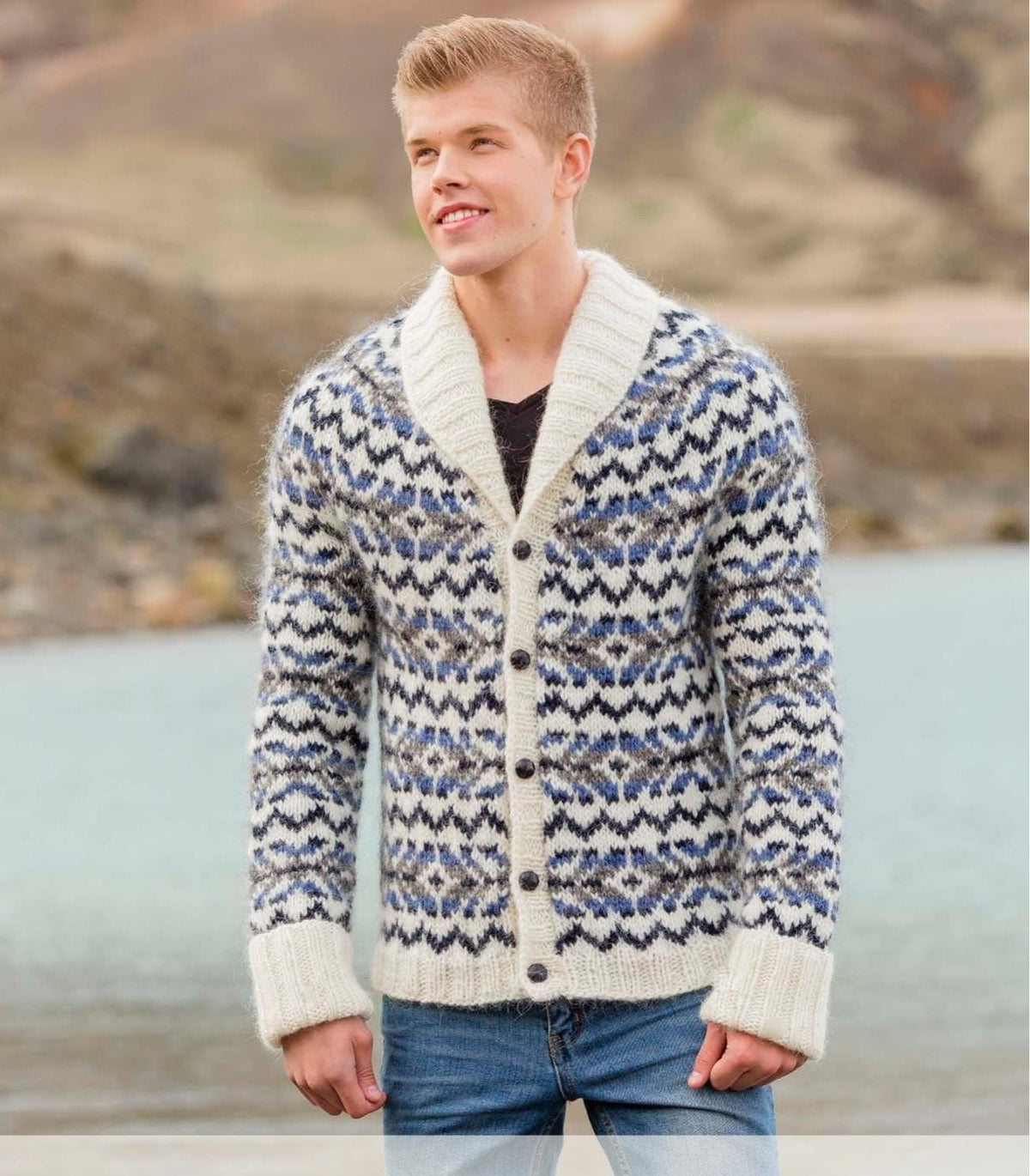 Young man by a lake in Rökkur from Lopi 33, shown in the pattern’s own colours — white Álafosslopi patterned all over with midnight blue zigzags and bands of denim heather, midnight blue and grey heather, a deep white ribbed shawl collar, a ribbed hem, long ribbed cuffs folded back and five dark buttons. The black T-shirt and the jeans are not part of the pattern