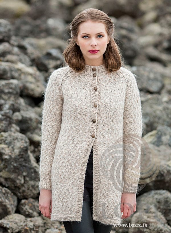 Woman in front of a wall of lava rocks in Þórunn — a long light beige cardigan textured all over with small slipped-stitch cables, raglan lines, a garter-stitch stand-up neckband, garter-stitch cuffs and front bands, and six buttons on the upper half, open below. The black top and the black trousers are not part of the pattern