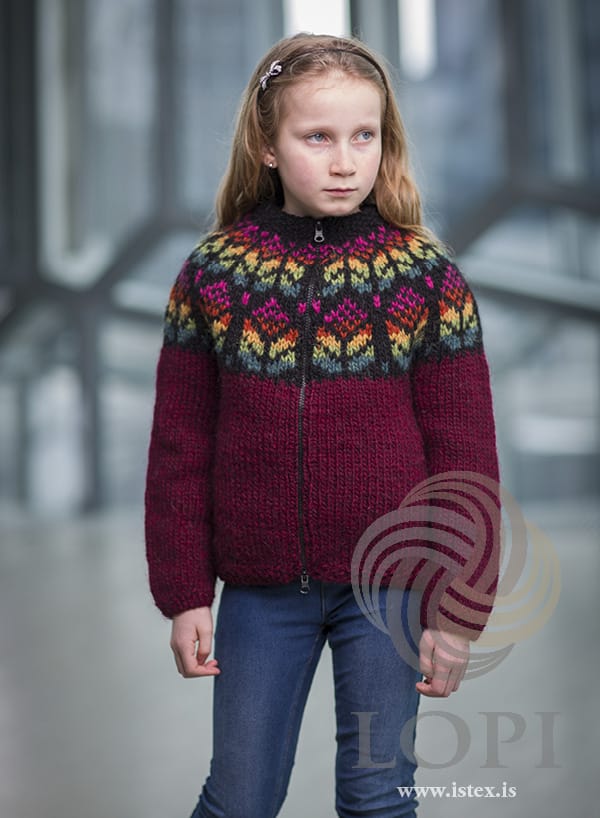 Girl in Blómey — an oxblood red Álafosslopi zip cardigan with moss-stitch edges, a black yoke with two rows of flowers shaded teal, green, gold, orange and magenta, and a black collar, shown in the pattern’s original colours. The jeans, the hairband and the earring are not part of the pattern