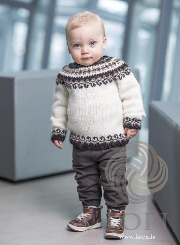 Toddler standing in a bright room in Bogi, shown in the pattern’s own colours — a white Léttlopi sweater with a round yoke of black sheep (dark brown) and murky (light brown) motifs, and matching bands above the black sheep moss-stitch hem and cuffs. The brown trousers and the trainers are not part of the pattern