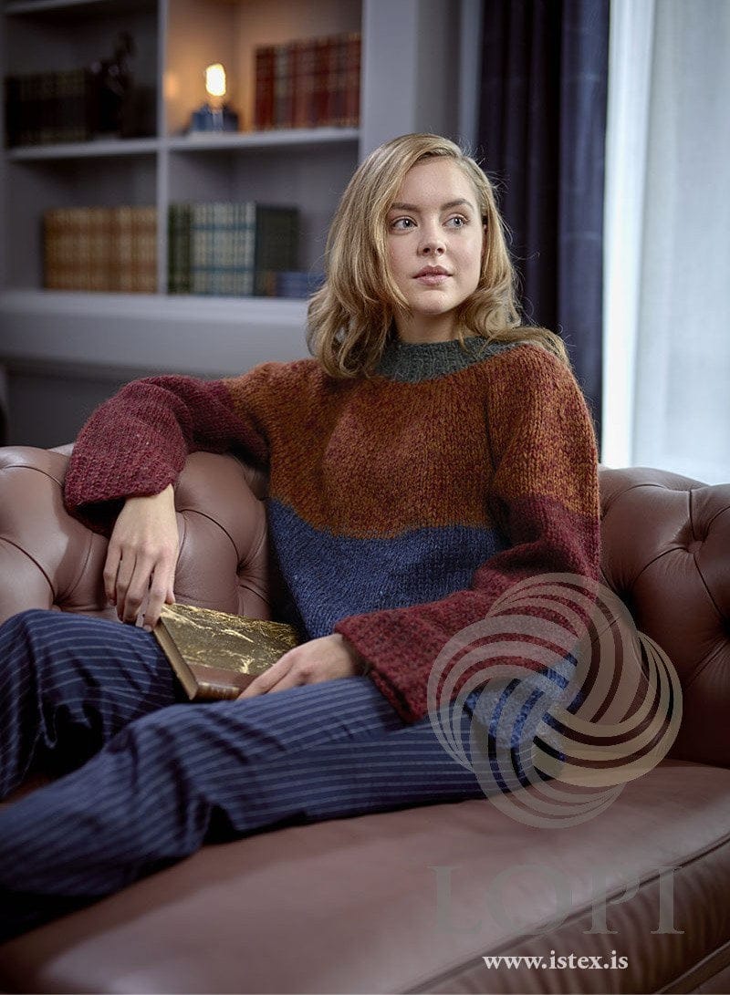 Woman seated on a leather sofa in the Fletir sweater — the rust-amber yoke, the deep red sleeves with their long cuffs, the blue lower body and the dark neckband (shown in the original colours — this kit has forest green in place of the forest in the neckband). The striped trousers and the book are not part of the pattern
