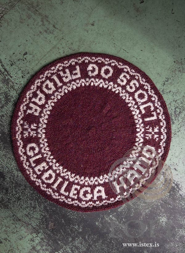 Friður round mat in ruby red Álafosslopi with an ecru border reading GLEÐILEGA HÁTÍÐ · LJÓSS OG FRIÐAR