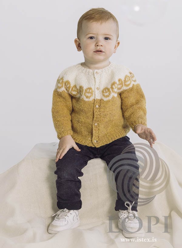 Toddler sitting on a cream cloth in Bros, seen from the front — a golden yellow baby cardigan with a white yoke of round yellow smiling faces above a wavy yellow edge, a white rib neckband and small round buttons, shown in the pattern’s own colours. The dark jeans, the white trainers, the cream cloth and the faint soap bubble at the top are not part of the pattern