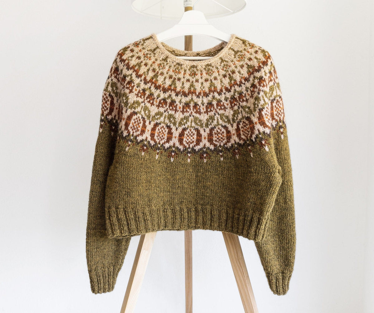 Pomegranate Lopapeysa on a hanger: olive-green golden heather body and long sleeves, and a straw yoke with rust pomegranates, olive leaves and a chocolate zigzag edge — a women’s L knitted cropped