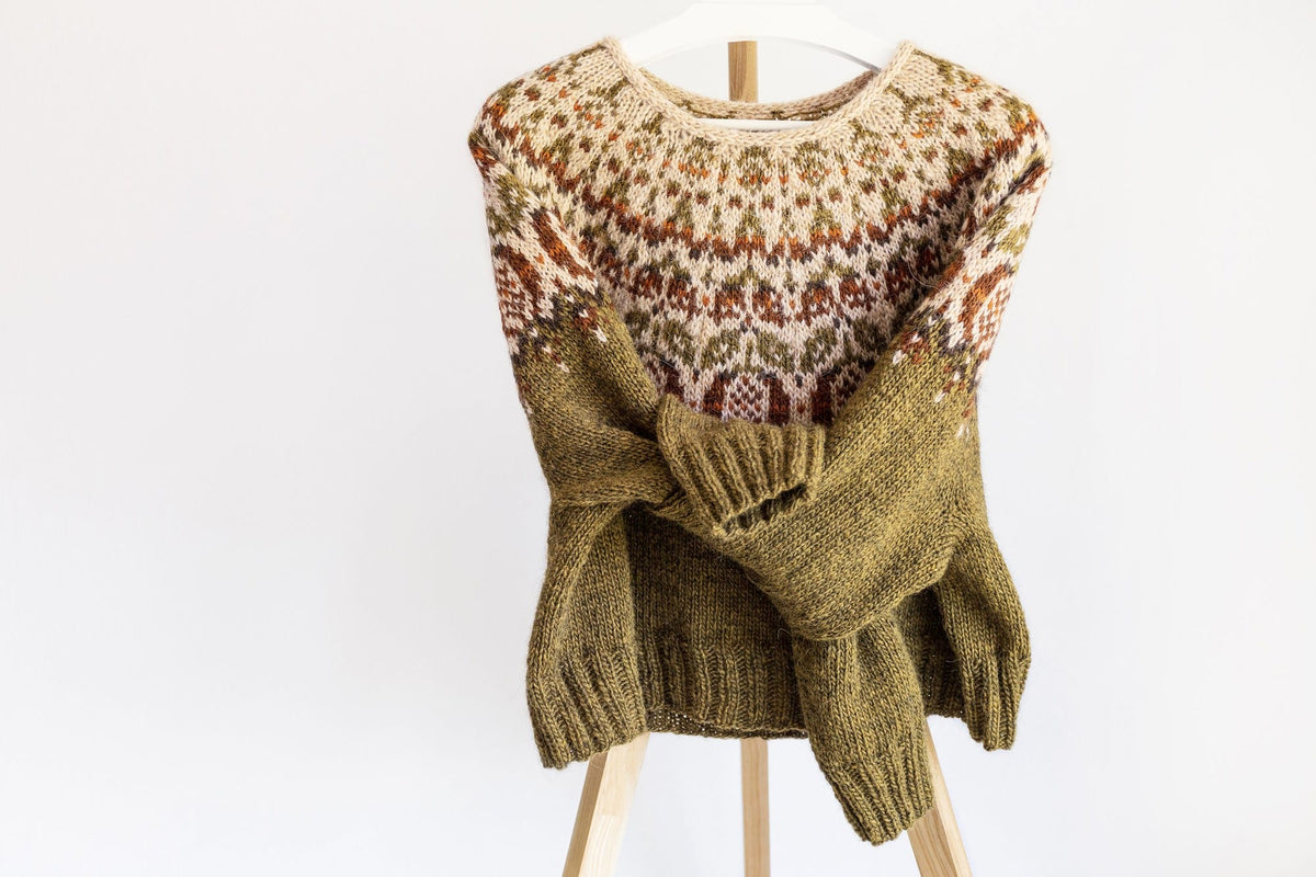 Pomegranate Lopapeysa on a wooden stand with the sleeves folded across the front, showing the k2, p2 rib cuffs and hem