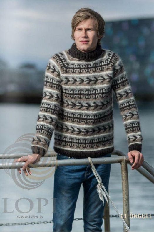 Young man at a metal railing by the water in Öngull, shown in the pattern’s own colours — a raglan pullover striped in white, oatmeal, light beige, light ash and ash heather, with bands of black-sheep arrows and hooks over the body and sleeves, black-sheep rib at the hem and cuffs and a tall black-sheep collar folded over. The jeans are not part of the pattern