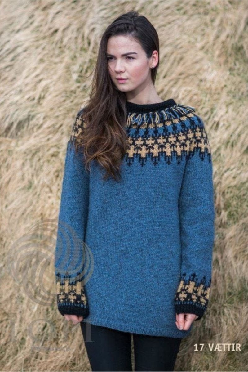 Woman in front of windblown dry grass in Vættir, shown in the pattern’s own colours — a long stone blue Léttlopi sweater with a yoke of camel crosses on black and a band of blue and black, a black collar with a fringe of blue, camel and black strands, camel-and-black borders above black rib cuffs, and a blue rib hem. The black trousers and the stud earring are not part of the pattern
