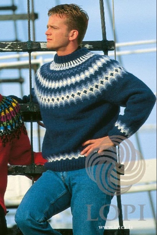 Man leaning on a rope ladder in Lopi 12 no. 19, the navy version, in the pattern’s own colours — navy with a yoke of white, light denim and denim bands with small navy crosses, a band of small diamonds and short white lines below the folded navy neckband, and a stepped band above the ribbed hem and cuffs. His blue jeans are not part of the pattern; the red cardigan at the left edge is Lopi 12 no. 17, a different pattern, and the black trousers below it are not part of either