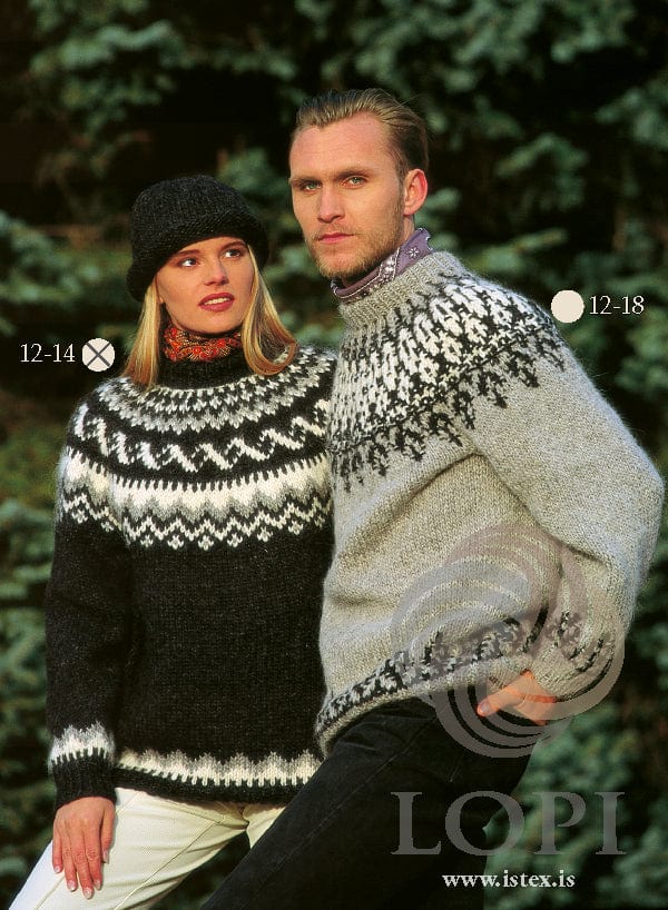 Blonde woman among evergreen trees in the Lopi 12 no. 14 sweater and the hat from page 3, in the pattern’s own colours — black heather with a round yoke of small white motifs, a light grey band zigzagging into white, slanting white waves and short white stripes, the border above the ribbed hem and cuffs, and a black heather hat with a rolled lower edge. Her red paisley scarf and cream trousers are not part of the pattern; the man (lilac collar, dark trousers) wears Lopi 12 no. 18, a different pattern