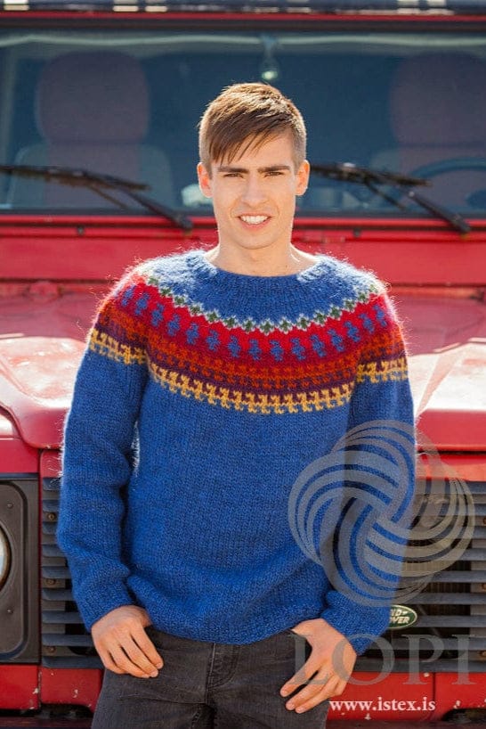 Young man standing in front of a red off-road vehicle wearing Ás in the pattern’s original colours — a space blue sweater with a round yoke of golden, gloxinia, burnt orange, dusk red and happy red bands, small space-blue shapes on the red, and garden-green and light-denim motifs near the neck. The dark jeans and the vehicle are not part of the pattern