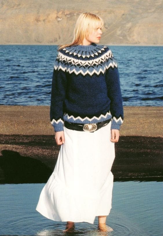 Blonde woman standing barefoot in shallow water, sea and hills behind, wearing the No. 5 design knitted in navy and a mid blue — the colours of our navy kit, not the pattern’s original greens — with white zigzags and black motifs, mid-blue rib and a mid-blue neckband. This photo is not in the pattern. The long white skirt and the black belt with a large silver buckle are not part of the pattern