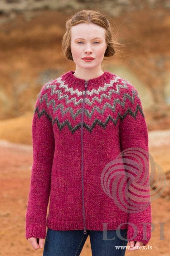 Young woman against ochre and red earth in the fuchsia Blær in the ladies’ style — a fuchsia Álafosslopi zip cardigan with plain body and sleeves and a yoke of zigzag bands in black and greys, lightest near the neck. This photo is from Ístex’s photo set for the pattern, not the PDF. The blue jeans are not part of the pattern