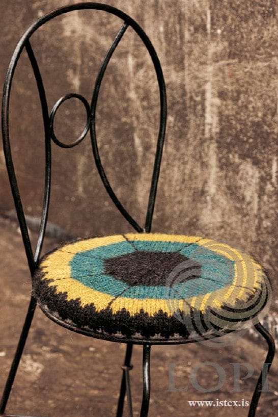 Sneið on the same kind of black wrought-iron chair, showing the green side in the pattern’s own colours — a black heather centre, rings of teal and chartreuse, dark backstitched lines and a black heather sawtooth border round the side. The chair is not part of the pattern