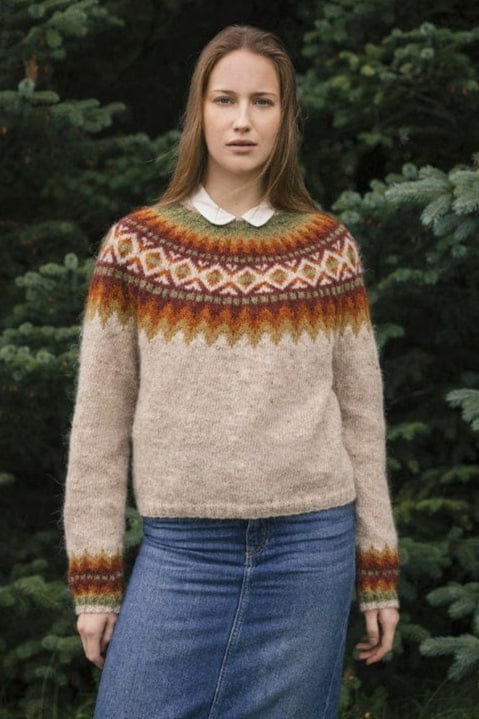 Young woman in front of spruce trees wearing Góa in the pattern’s own colours — a light moorit sweater that ends at the waist, with a round yoke of mustard and ginger zigzags, marron, moss and light diamonds, and matching bands above the moss rib cuffs. The white blouse with a round collar under it and the long denim skirt are not part of the pattern