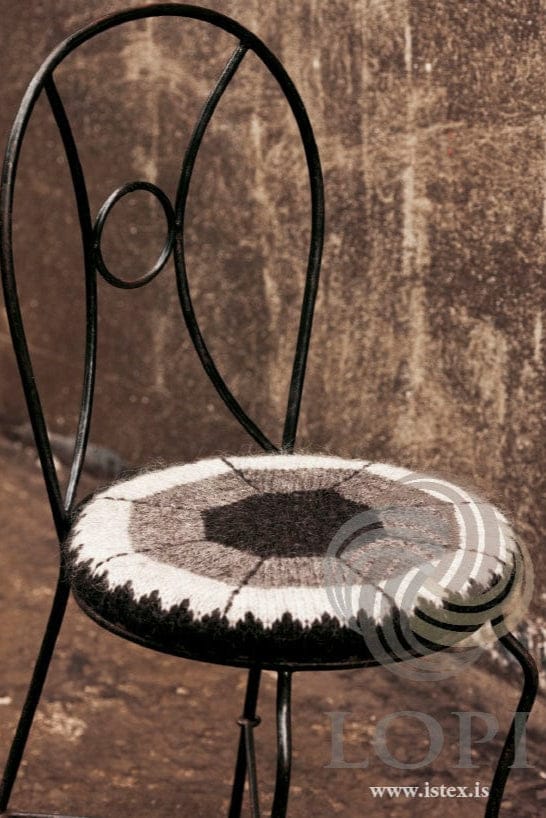 Sneið on the round seat of a black wrought-iron chair against a weathered wall, showing the grey side in the pattern’s own colours — a black heather octagon in the centre, rings of grey and ash, dark backstitched lines running to the edge, and a small black heather sawtooth border round the side. The chair is not part of the pattern