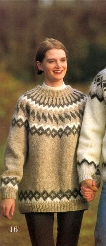 Smiling woman in the Lopi 12 no. 16 sweater, marked 16, in the pattern’s own colours — light beige heather with a yoke of light beige diamonds in dark grey with white points above, slanting dark grey diamonds, a white and dark grey zigzag and fine dark grey lines up to the neckband, and the diamond band above the ribbed hem and cuffs. Her white polo neck and dark tights or leggings are not part of the pattern, nor is the man holding her hand on the right, in another pattern’s white sweater and blue jeans