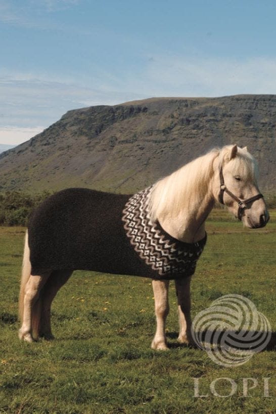 A pale cream Icelandic horse with a white mane in a grassy field, a steep mountain behind, wearing Hestur in the pattern’s own colours — a black heather garter-stitch blanket over its back and the patterned yoke round its neck and chest, with zigzag bands of oatmeal and white and a row of white diamonds. The halter is not part of the pattern; the LOPI logo is printed in the corner