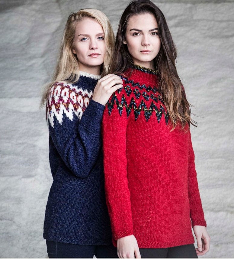 Two women in the two Jökla colourways, shown in the pattern’s own colours — on the left the blue one, navy with a white zigzag yoke, white ovals in a flecked red band and a white collar edge; on the right the red one, crimson with a black multicoloured zigzag-and-drop yoke and a flecked collar edge. The dark trousers and the earring are not part of the pattern
