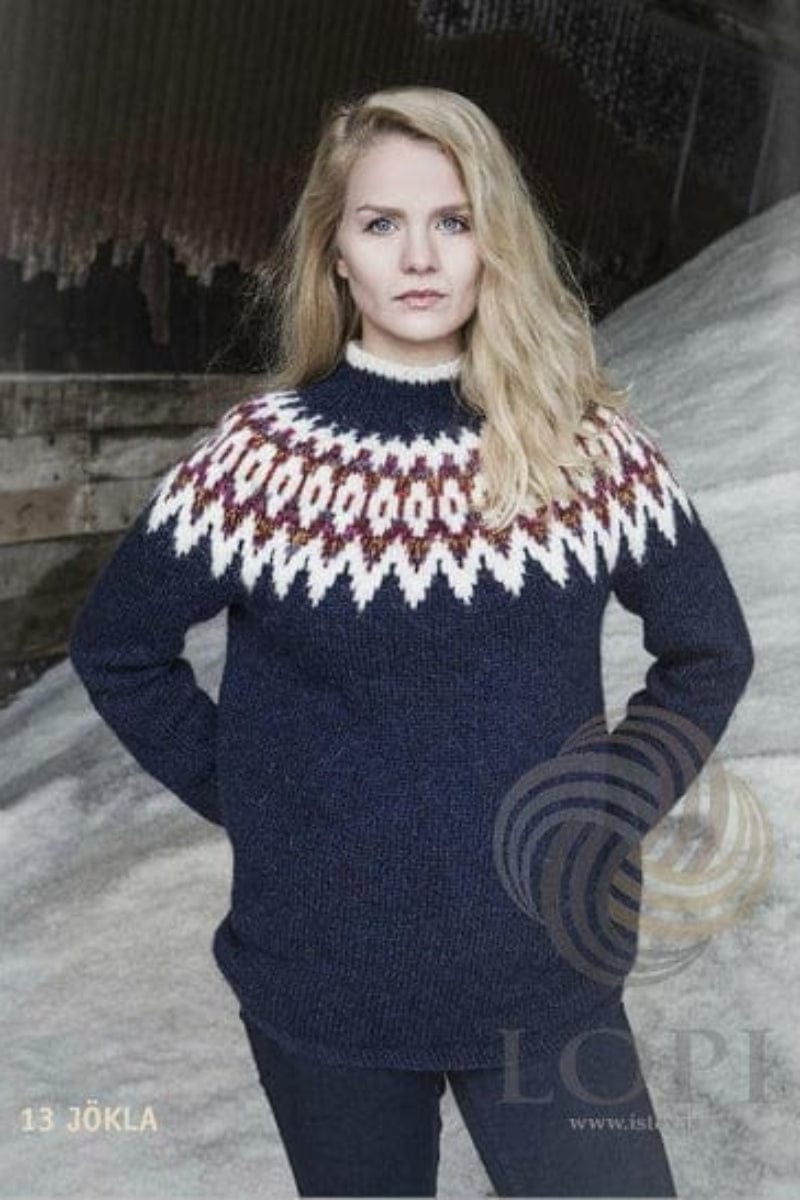 The blue Jökla, shown in the pattern’s own colours — navy blue Léttlopi with a white zigzag yoke, white ovals in a flecked red band, moss-stitch edges and a white collar edge. The dark jeans are not part of the pattern