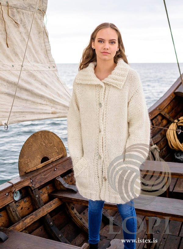 Young woman on the deck of a wooden boat in front of a sail, wearing Júní buttoned with both hands in the pockets — a long, loose white cardigan with a deep ribbed collar folded over and textured rib at the hem, cuffs and front bands, as photographed for the pattern. The blue jeans and the earrings are not part of the pattern