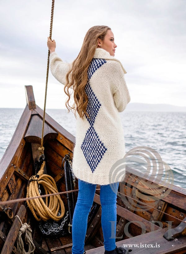 Júní from the back at the bow of a wooden boat: a column of navy-and-white diamonds runs up the centre back of the long white cardigan, from the rib to the collar, as photographed for the pattern. The blue jeans and the earrings are not part of the pattern